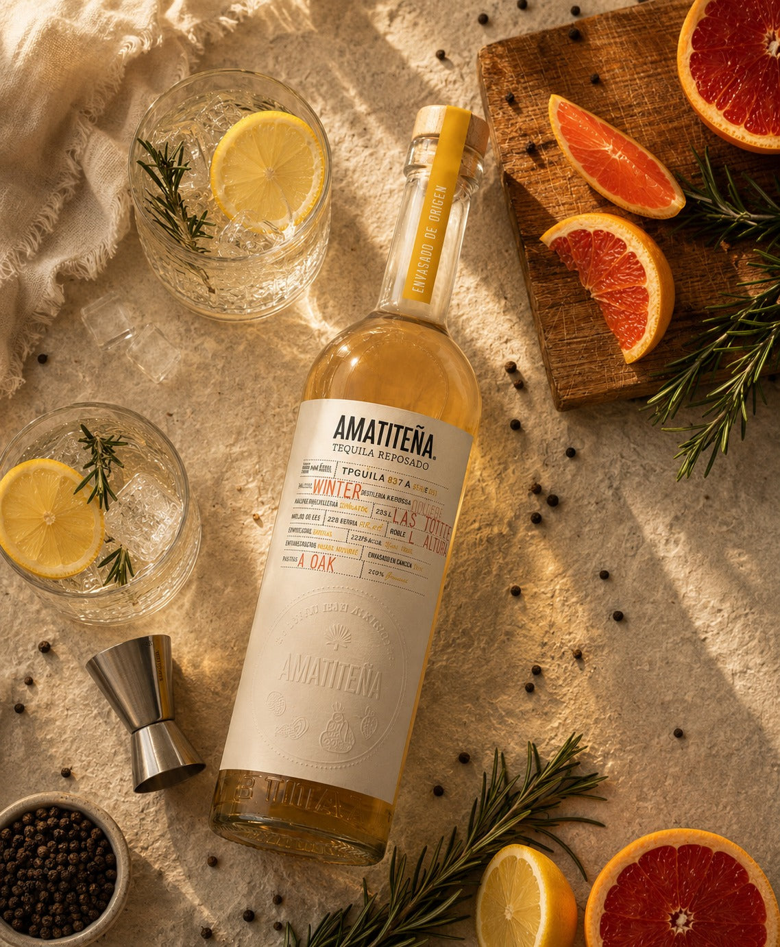 A bottle of Amatteña Trujillo Single Malto Tequila Blanco is styled in a warm, rustic flat-lay composition with cocktail glasses, citrus slices, rosemary, and spices on a textured beige fabric background.