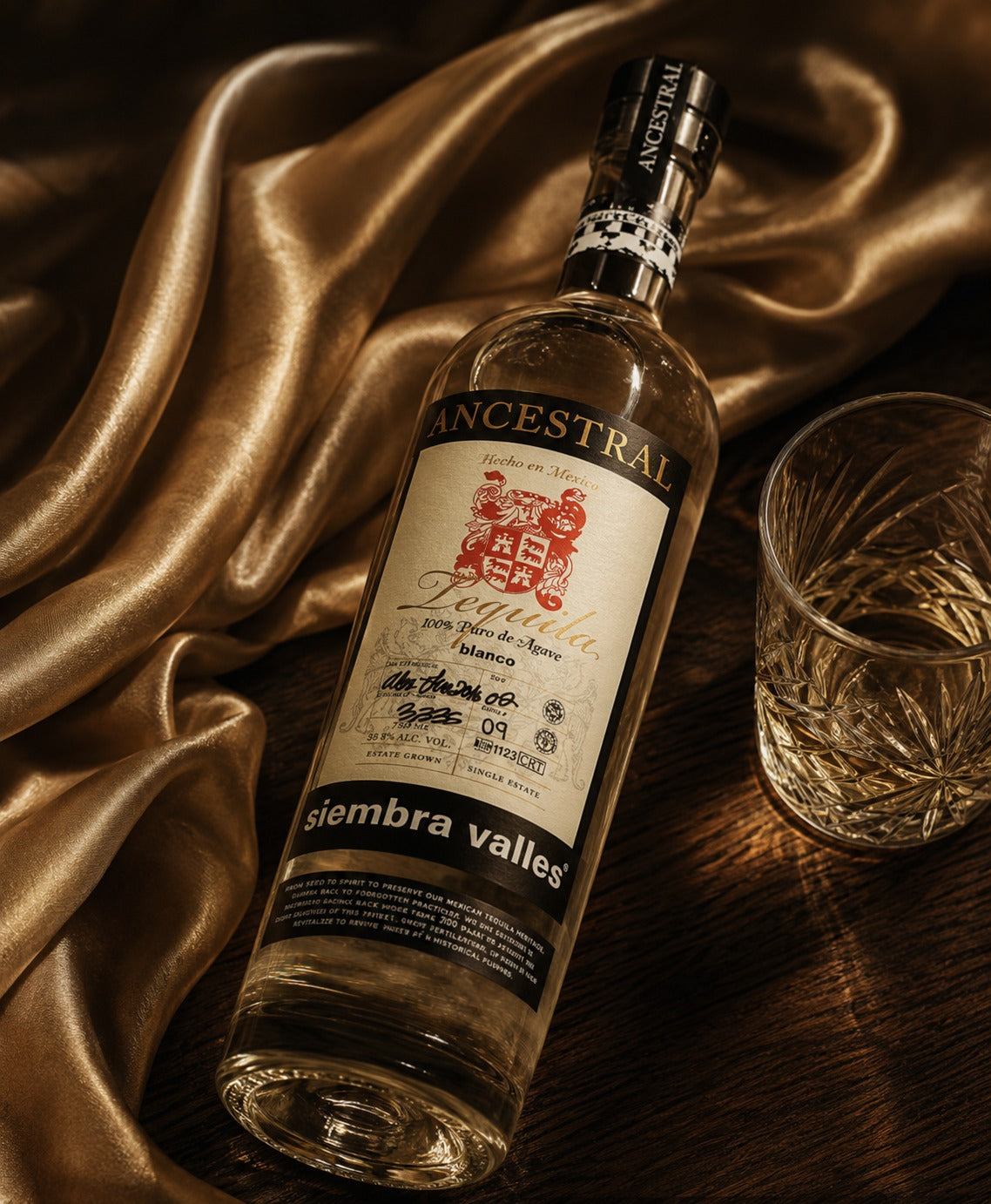 A bottle of Ancestral Sierra Valles Blanco tequila is displayed diagonally on a dark wooden surface next to a cut crystal glass, draped with elegant gold satin fabric.