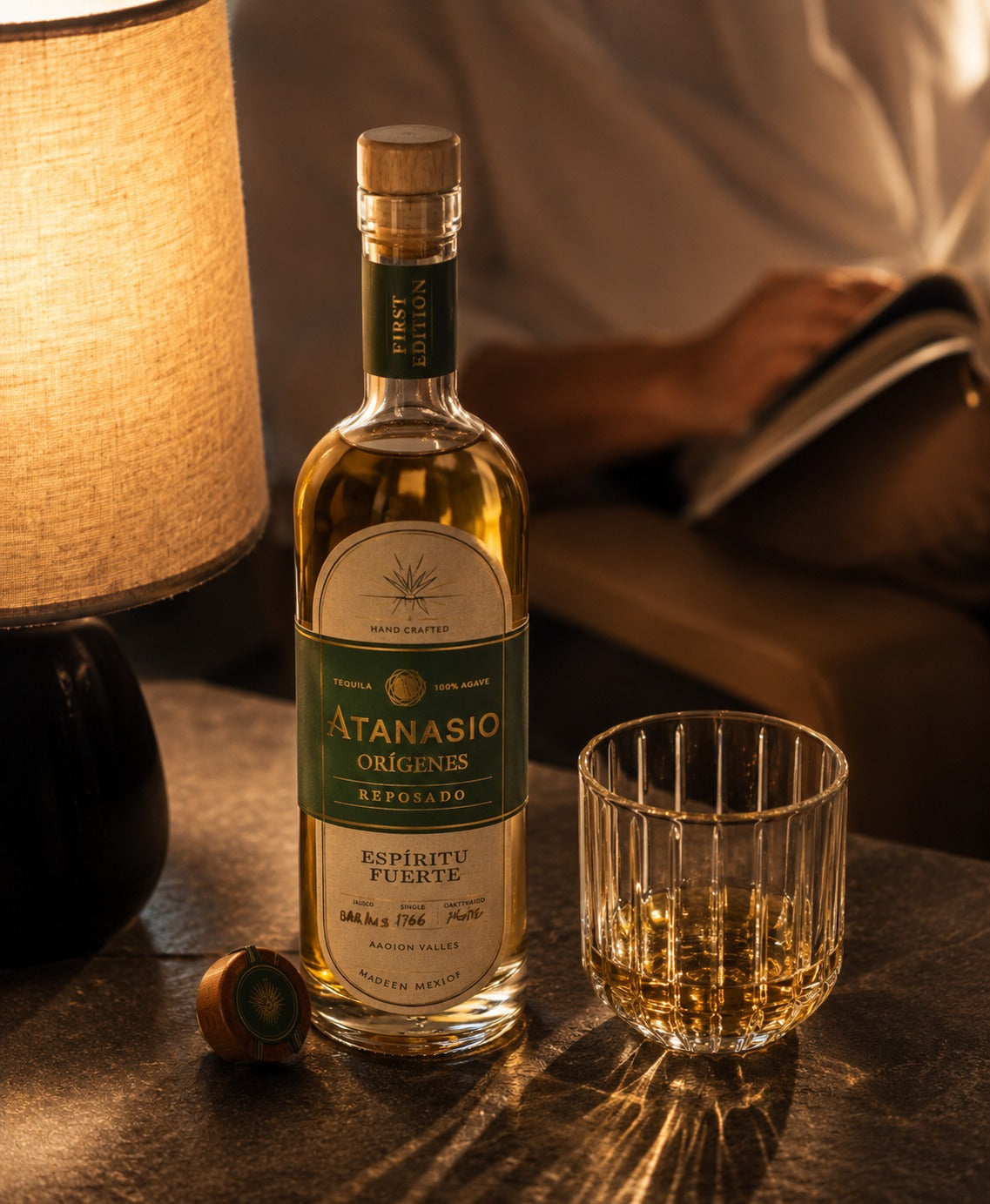 A bottle of Atanasio Espiritu Fuerte rum with its cap off, placed on a dark table next to a filled drinking glass and a lit lamp, with a person reading in the background.