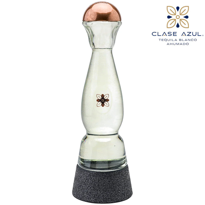A clear glass tequila decanter with a copper-colored top, a small black floral emblem on the front, and a textured dark gray base. The Clase Azul logo is visible in the upper right corner against a plain white background.