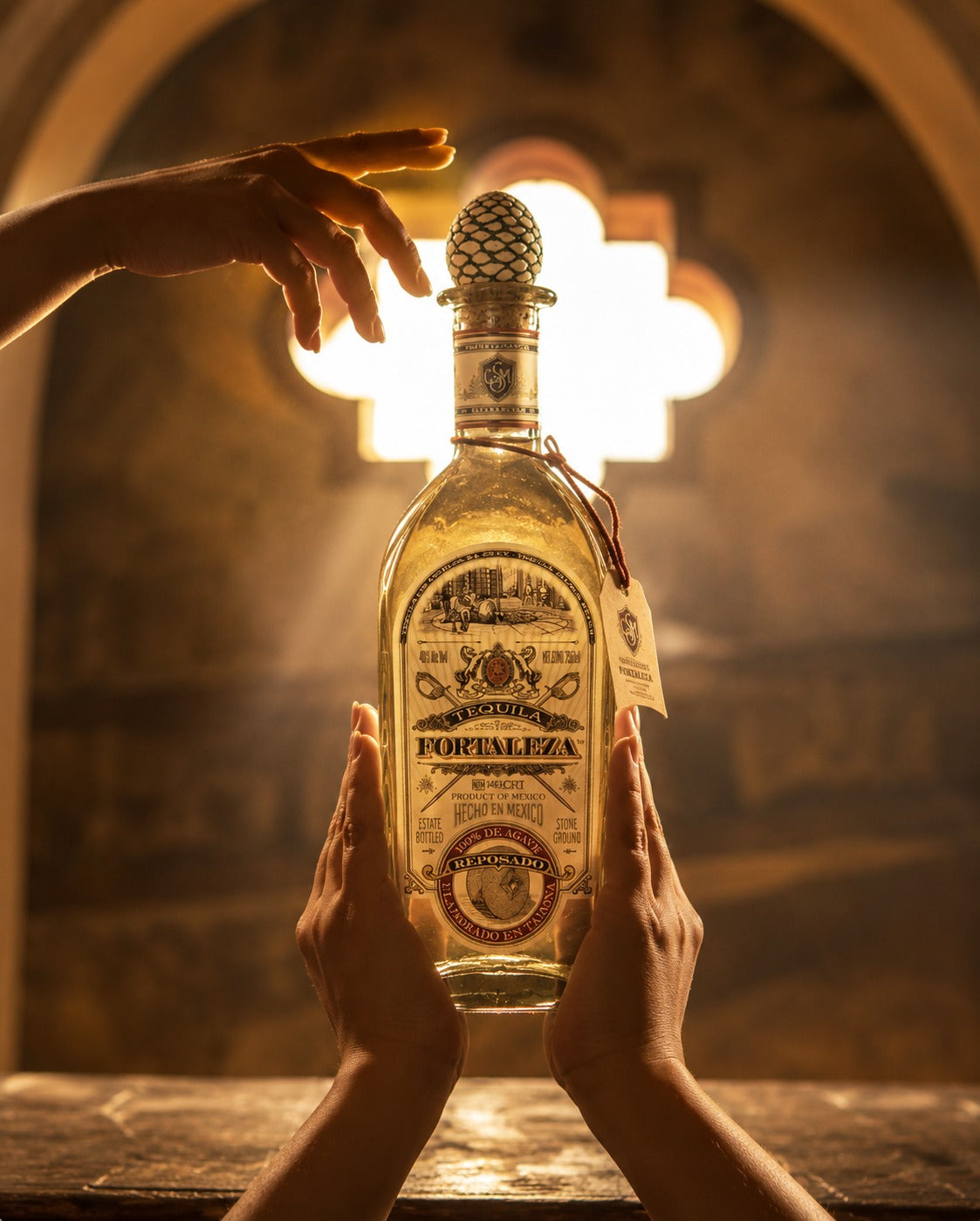 A bottle of Forajaleza tequila is held by two hands in front of a warm, glowing arched background. A third hand reaches from the top left toward the bottle.
