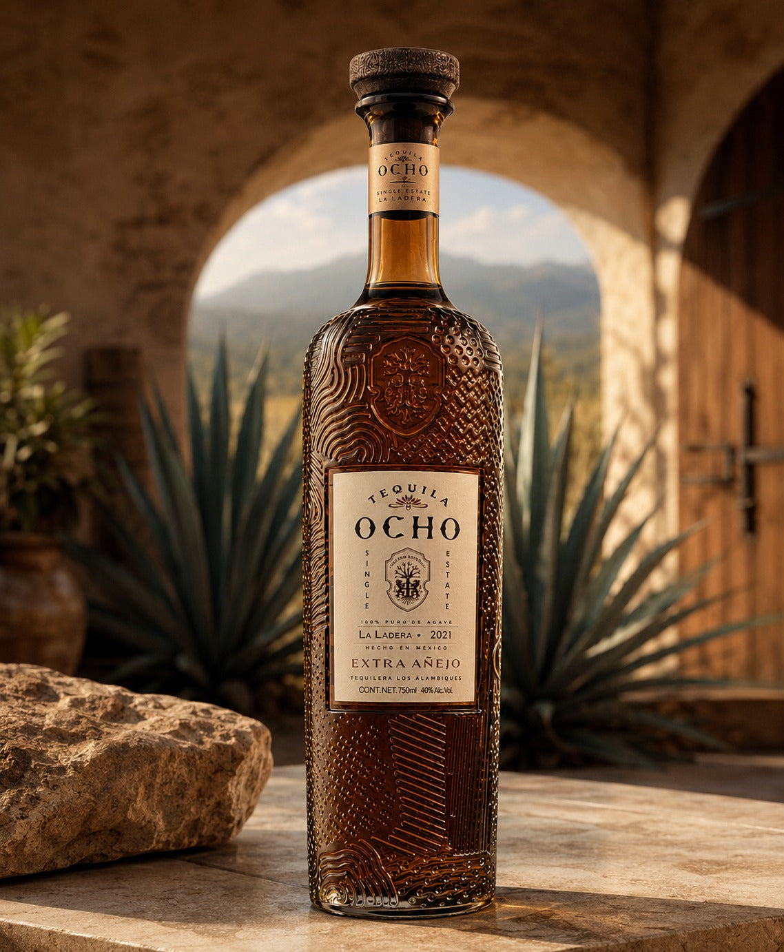 A bottle of Ocho Tequila Extra Añejo tequila stands on a stone surface in front of an arched doorway with agave plants and mountains in the background. The bottle has a textured dark brown glass body, a cream label, and a dark cork stopper.