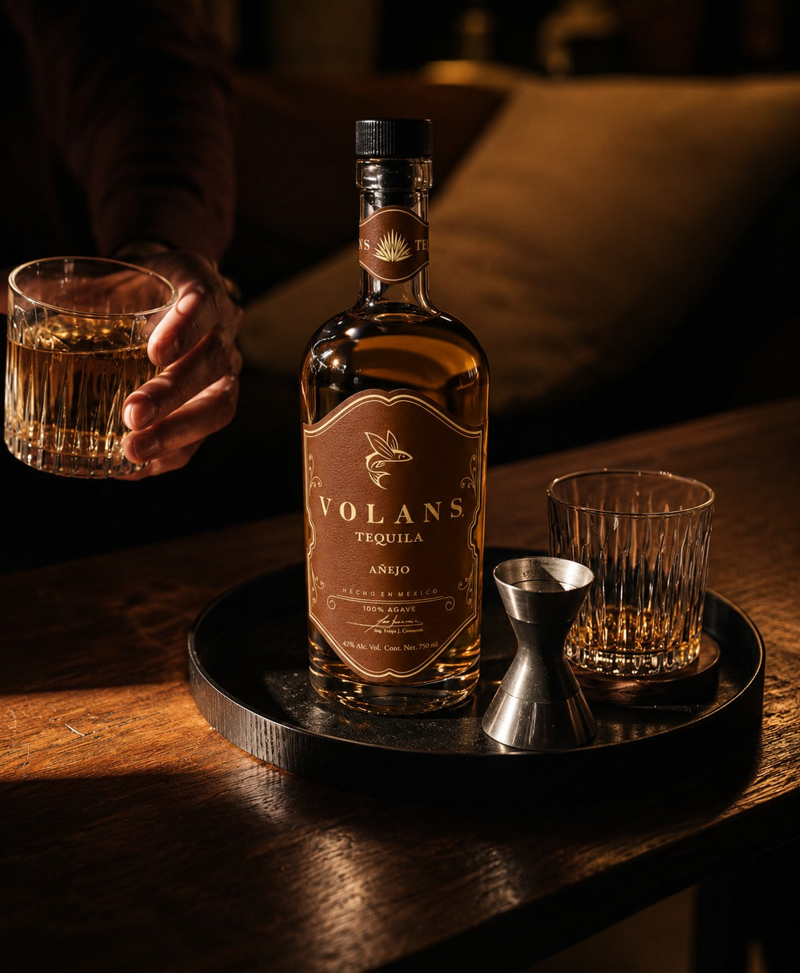 A bottle of Volans Tequila AS120 stands on a dark tray on a wooden table, with two glasses and a metal jigger beside it. A hand holding a filled glass is visible at the left edge in warm, moody lighting.
